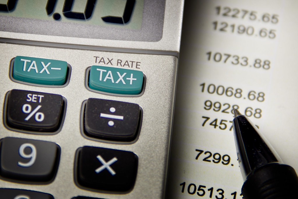 TAX+·TAX- 버튼이 있는 계산기와 숫자가 인쇄된 명세서 위에 놓인 펜 클로즈업 — 세금 계산 장면